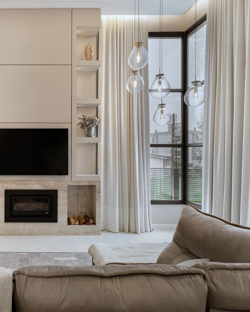 Sala de estar com boiserie clara, painel ripado de madeira, lareira revestida em mármore e cortina alta filtrando a luz