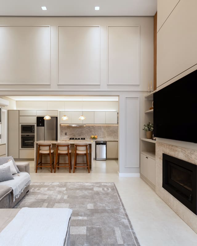 Sala de estar com boiserie clara, lareira revestida em mármore e vista para a cozinha com ilha e banquetas de madeira